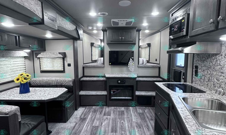 Interior view of a modern RV showing a kitchen with double sinks, cabinets, and a microwave on the right, and a seating area with a table on the left