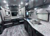 Interior of a modern RV kitchen and living area with dark cabinets, island sink, microwave, and stairs to seating area.
