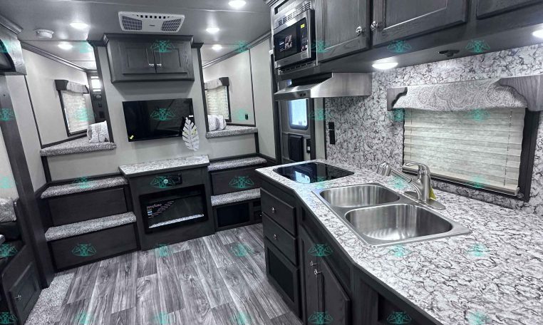 Interior of a modern RV kitchen and living area with dark cabinets, island sink, microwave, and stairs to seating area.
