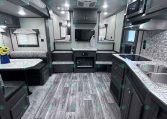 Interior of a modern travel trailer: kitchen on the right with sink, counters, and cabinets, opposite seating area with a TV center.