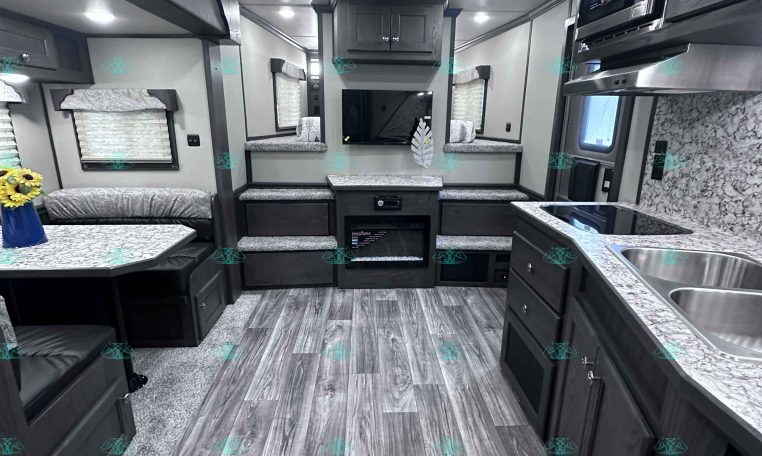Interior of a modern travel trailer: kitchen on the right with sink, counters, and cabinets, opposite seating area with a TV center.