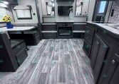 Interior of a modern RV/trailer living area with built-in dinette, couch seating, and entertainment center against a wall-mounted TV in the center.