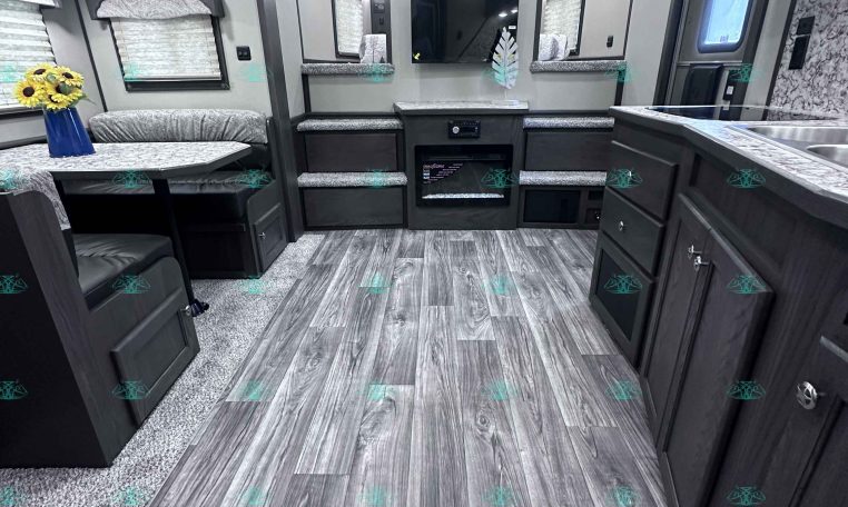 Interior of a modern RV/trailer living area with built-in dinette, couch seating, and entertainment center against a wall-mounted TV in the center.