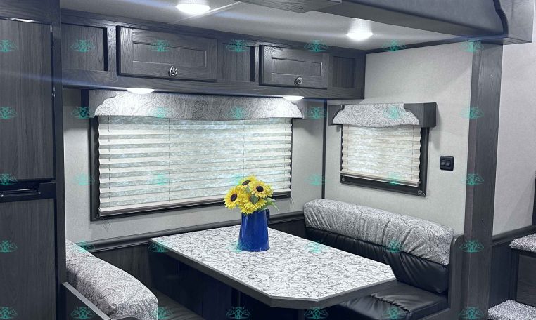 RV dining nook with dark wood booths, patterned cushions, and a marble-pattern table beneath overhead cabinets and a decorative valance without natural light sources visible