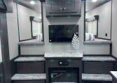 Interior of a modern trailer living area with dark wood cabinets above a wall-mounted TV, granite counters, and a small electric fireplace beneath.