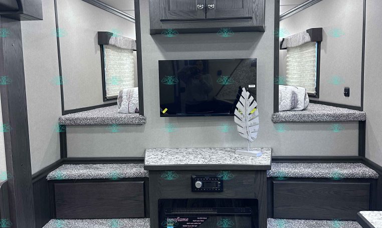 Interior of a modern trailer living area with dark wood cabinets above a wall-mounted TV, granite counters, and a small electric fireplace beneath.
