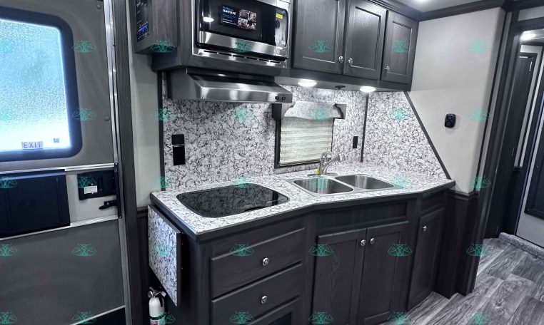 RV kitchen with dark wood cabinets, stainless microwave, stovetop, and double sink on a marble backsplash counter area