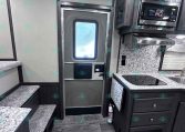Interior of a modern RV kitchen with stainless steel microwave, vent hood, marble counters, and dark wood cabinets; stairs on left and outside door ahead.