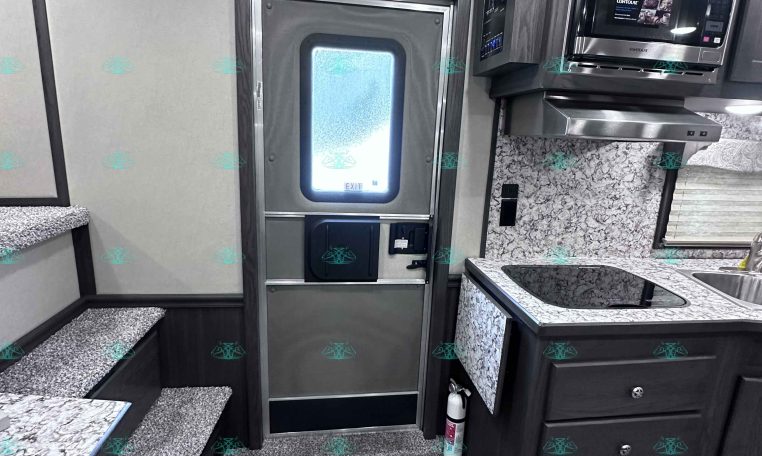 Interior of a modern RV kitchen with stainless steel microwave, vent hood, marble counters, and dark wood cabinets; stairs on left and outside door ahead.