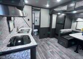 Interior of a modern travel trailer with a kitchen island, double sink, and stove along a marble countertop wall; living/dining area ahead with a booth-style dinette and dark wood cabinetry.