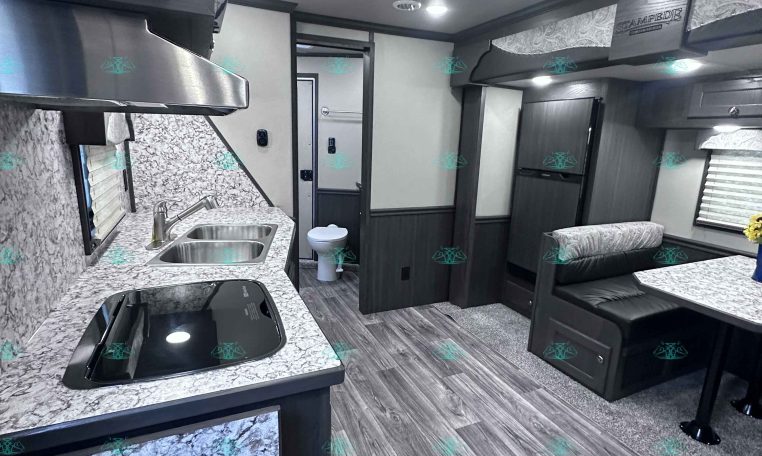 Interior of a modern travel trailer with a kitchen island, double sink, and stove along a marble countertop wall; living/dining area ahead with a booth-style dinette and dark wood cabinetry.