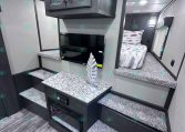 Compact RV interior with dark wood upper cabinet, wall-mounted TV, granite-look countertop, and steps leading to a bedroom with patterned bedding in the back.