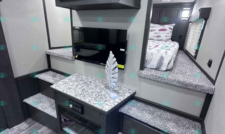 Compact RV interior with dark wood upper cabinet, wall-mounted TV, granite-look countertop, and steps leading to a bedroom with patterned bedding in the back.