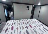 Interior bedroom of a travel trailer with a feather-pattern quilted bedspread and overhead vents/speaker on the ceiling, gray walls framing a doorway.