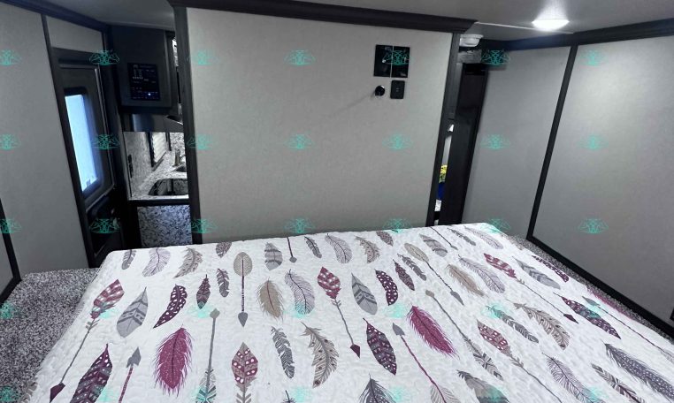 Interior bedroom of a travel trailer with a feather-pattern quilted bedspread and overhead vents/speaker on the ceiling, gray walls framing a doorway.