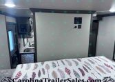 RV bedroom with a feather-patterned quilted bed in the foreground and a kitchenette visible to the left.