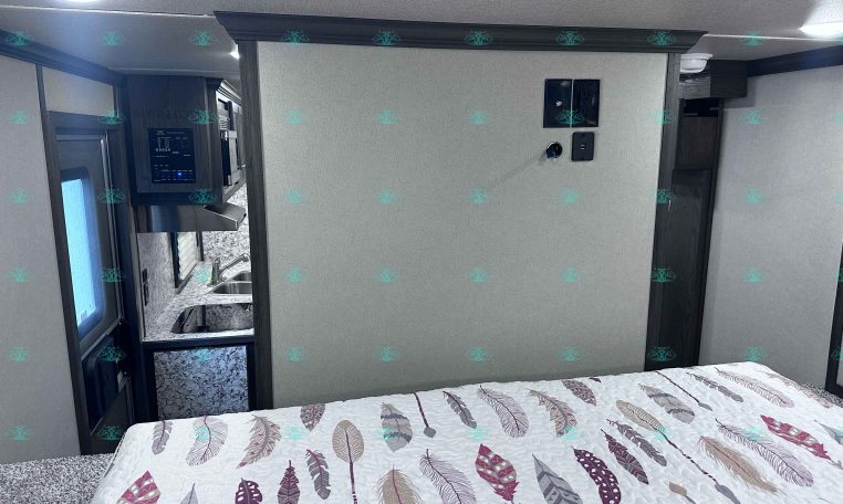 RV bedroom with a feather-patterned quilted bed in the foreground and a kitchenette visible to the left.