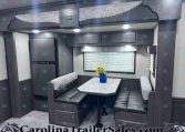 Dark wood RV dining area with U-shaped booth seating around a marble table, upper cabinets, window with blinds, and a blue vase of sunflowers on the table.