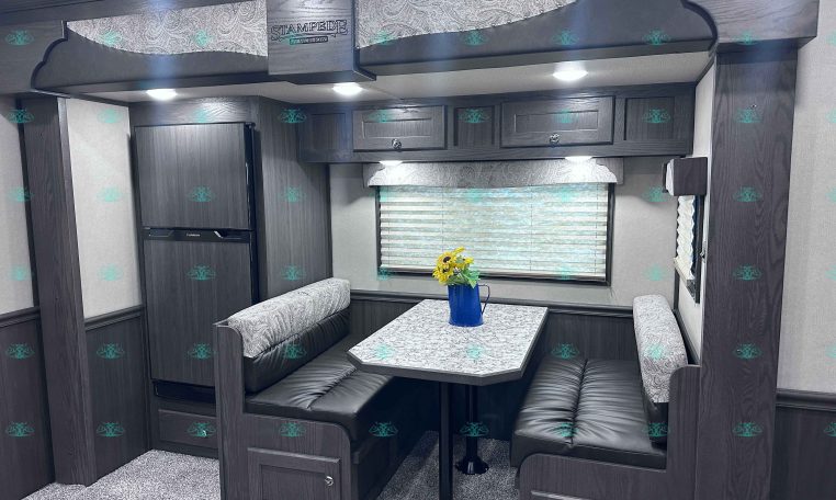Dark wood RV dining area with U-shaped booth seating around a marble table, upper cabinets, window with blinds, and a blue vase of sunflowers on the table.