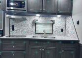 Dark wood kitchen cabinets with built-in microwave and range hood, marble backsplash, and double sink under a window in a trailer kitchen.