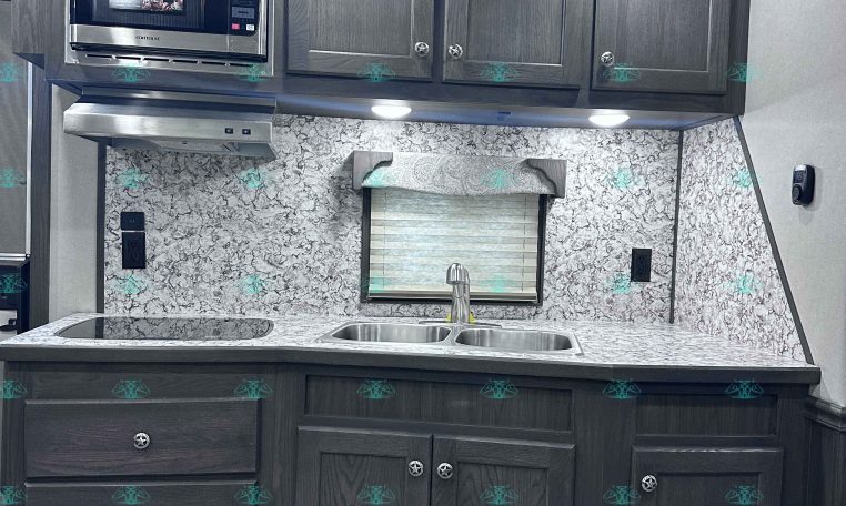 Dark wood kitchen cabinets with built-in microwave and range hood, marble backsplash, and double sink under a window in a trailer kitchen.