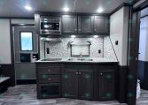 Interior of a modern trailer kitchen with dark wood cabinets, marble backsplash, double sink, and stainless microwave above the stove area; ceiling lights illuminate the space.