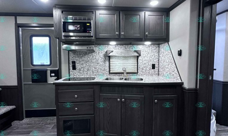 Interior of a modern trailer kitchen with dark wood cabinets, marble backsplash, double sink, and stainless microwave above the stove area; ceiling lights illuminate the space.