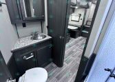RV interior showing bathroom with sink and granite counter, mirrored medicine cabinet, and adjacent toilet; doorway leads to a bedroom with bunks in a dark wood finish, gray walls, and wood-look flooring.
