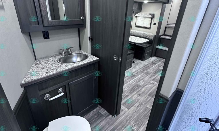 RV interior showing bathroom with sink and granite counter, mirrored medicine cabinet, and adjacent toilet; doorway leads to a bedroom with bunks in a dark wood finish, gray walls, and wood-look flooring.