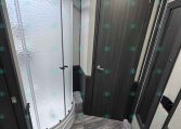 Compact trailer bathroom with curved frosted glass shower, dark wood storage cabinet, and a gray-toned vanity door.