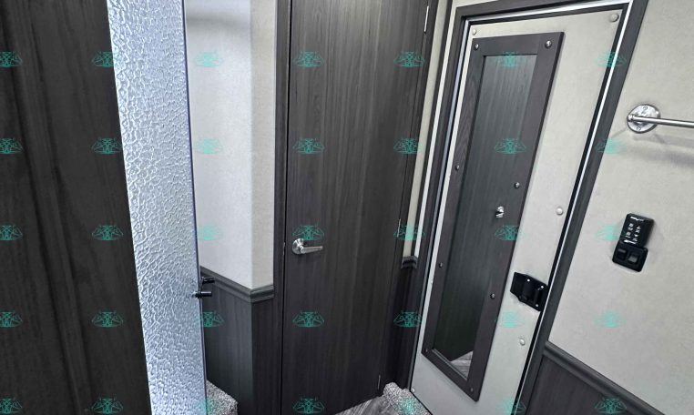 Compact trailer bathroom with a curved glass shower door on the left, dark wood cabinet doors, and a seated toilet on the right.