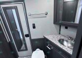 Compact trailer bathroom with toilet, marble countertop sink, and dark wood cabinets with a wall mirror.