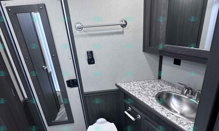 Compact trailer bathroom with toilet, marble countertop sink, and dark wood cabinets with a wall mirror.