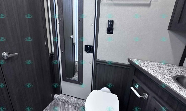 RV bathroom with a white toilet, granite countertop, and dark wood cabinets; a towel bar on the wall and a slim mirror cabinet nearby.