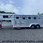Silver aluminum horse trailer with multiple windows, parked on a gravel lot; trees in the background and red equipment in the foreground.