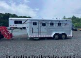 Silver aluminum horse trailer with multiple windows, parked on a gravel lot; trees in the background and red equipment in the foreground.