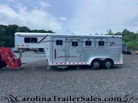 Silver aluminum horse trailer with multiple windows, parked on a gravel lot; trees in the background and red equipment in the foreground.