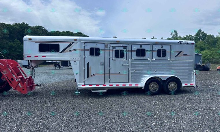 Silver aluminum horse trailer with multiple windows, parked on a gravel lot; trees in the background and red equipment in the foreground.