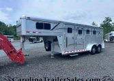 Silver aluminum gooseneck horse trailer with multiple stalls and windows, parked on a gravel lot with trees in the background.
