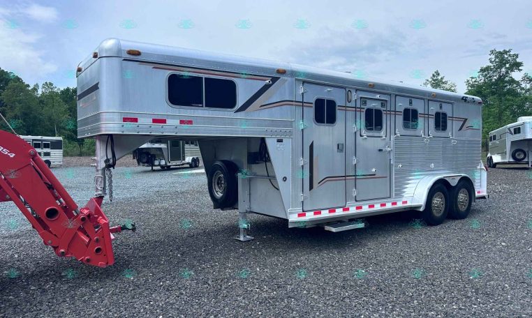 Silver aluminum gooseneck horse trailer with multiple stalls and windows, parked on a gravel lot with trees in the background.