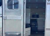 Rear view of an aluminum travel trailer with its door open, revealing a small kitchenette: fridge, microwave, and upper cabinets inside.