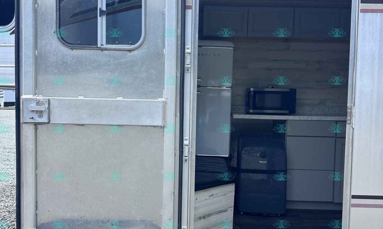 Rear view of an aluminum travel trailer with its door open, revealing a small kitchenette: fridge, microwave, and upper cabinets inside.