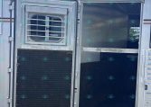 Open trailer door showing interior and a small barred window on the left door; metallic exterior with red/white reflective strip at bottom edge.
