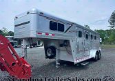 Silver gooseneck horse trailer with living quarters on a gravel lot, with red construction equipment in the foreground and trees in the background; CarolinaTrailerSales.com watermark visible.