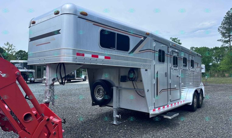 Silver gooseneck horse trailer with living quarters on a gravel lot, with red construction equipment in the foreground and trees in the background; CarolinaTrailerSales.com watermark visible.