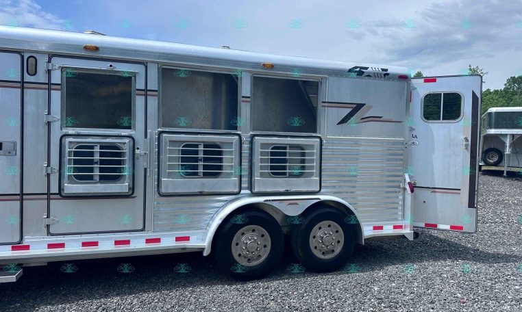 Silver aluminum horse trailer on a gravel lot with open stall doors and windows; another trailer is visible in the background.