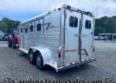 Silver aluminum horse trailer hitched to a red tractor in a gravel lot with trees and other trailers in the background, viewed from the side and rear.