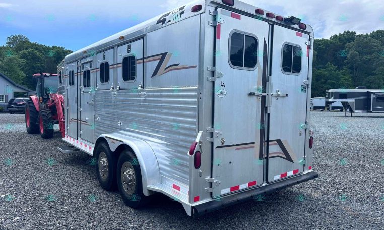 Silver aluminum horse trailer hitched to a red tractor in a gravel lot with trees and other trailers in the background, viewed from the side and rear.