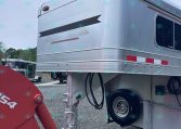 Front corner of a silver aluminum semi-trailer with rounded top, amber lights, and black stripe decals on a gravel lot.