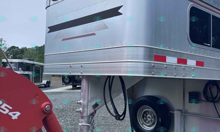 Front corner of a silver aluminum semi-trailer with rounded top, amber lights, and black stripe decals on a gravel lot.